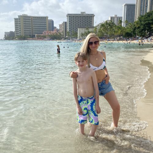 A blonde woman in a white bikini and jean shorts with her arm around a blonde boy with colorful swimming trunks, standing in the shallow ocean with a city behind them